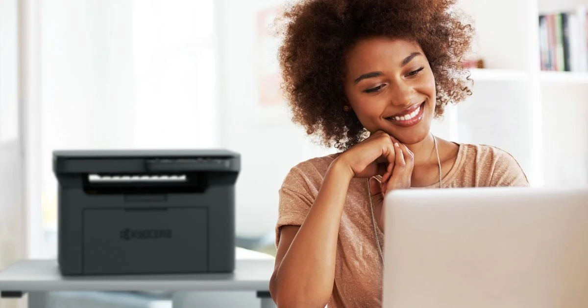 Smiling woman working on laptop with a Kyocera printer in the background.